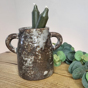 May include: A brown and white speckled ceramic vase with two handles. The vase is filled with two green candles.