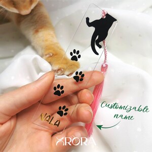May include: Clear acrylic bookmark with a black silhouette of a cat and paw prints. The bookmark has a pink tassel and a gold inscription that says "Nala". The text "Customizable name" is written on the image.