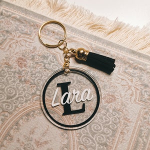 May include: A round, clear acrylic keyring with a black border and the name "Lara" written in white with a large black "L". The keyring has a gold chain and a black tassel.