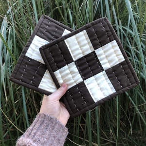K&ouml;nnte beinhalten: Zwei gequiltete Topflappen in Braun und Wei&szlig; mit Karomuster. Die Topflappen sind aus Stoff und haben ein gestepptes Design.