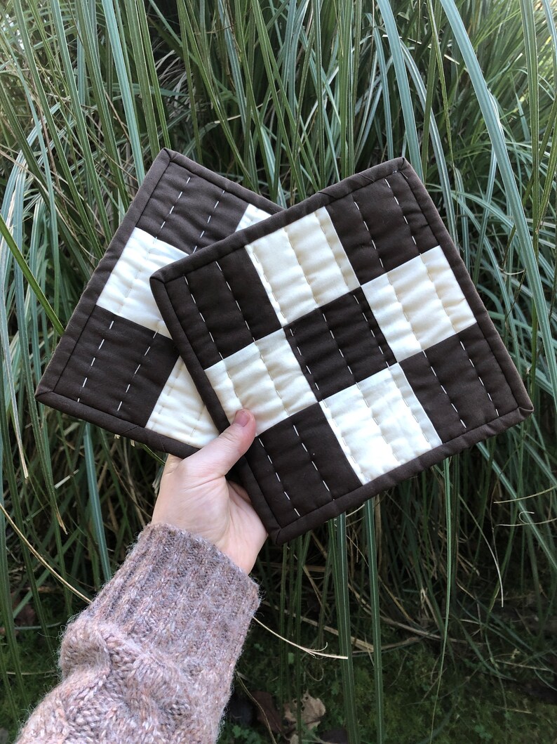 K&ouml;nnte beinhalten: Zwei gequiltete Topflappen in Braun und Wei&szlig; mit Karomuster. Die Topflappen sind aus Stoff und haben ein gestepptes Design.