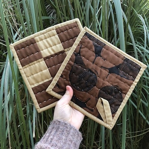 Puede incluir: Dos agarraderas de olla acolchadas. Una es marrón y beige con un patrón de cuadros. La otra es marrón con un patrón de caracoles estilizados.