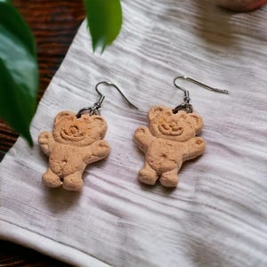 May include: A pair of light brown teddy bear earrings with silver ear wires. The earrings are made of a textured material and are displayed on a white cloth.