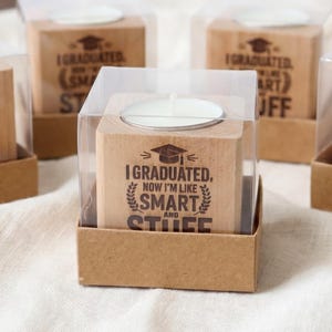 May include: Graduation-themed candle holders with clear plastic covers. The wooden blocks are printed with the text "I GRADUATED, NOW I'M LIKE SMART STUFF" and a graduation cap. A miniature graduation cap and a rolled diploma are in the background.