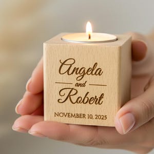 Può includere: Una candela accesa è posta su un piccolo blocco di legno quadrato. Il blocco è inciso con i nomi "Angela e Robert" e la data "10 novembre 2025". Il portacandele è tenuto tra le mani.