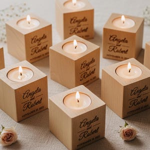 May include: Nine wooden candle holders with lit candles, arranged on a light-coloured surface. Each cube-shaped holder is engraved with the names "Angela and Robert" and a date. Small flowers and sprigs of greenery are scattered around the candles.