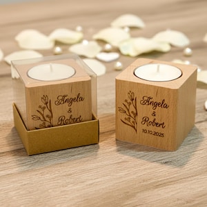 May include: Two wooden candle holders with tea lights, engraved with "Angela & Robert" and the date "10.10.2025". One is in a clear box. Floral design is engraved on the wood. White petals and pearls are scattered around.