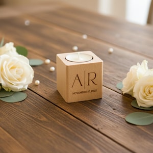 May include: A wooden candle holder with initials "AR" and the date "November 10, 2025" engraved on the front. The candle holder is surrounded by white roses, green leaves, and scattered pearls on a wooden table.