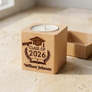 May include: A wooden candle holder with a graduation theme. It's engraved with "Class of 2026," a graduation cap, a diploma, and the name "William Johnson." A clear plastic box sits beside the candle holder.
