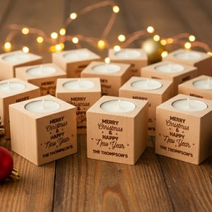 May include: A collection of wooden cube candle holders with tea lights, inscribed with "MERRY Christmas & HAPPY New Year" and "THE THOMPSON'S". The festive arrangement includes pine cones, red and gold baubles, and fairy lights, perfect for the holiday season.