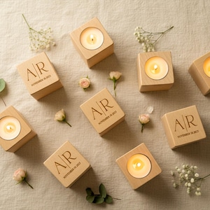May include: Wooden candle holders with lit candles, arranged on a neutral surface. Each holder is engraved with the initials "AR" and the date "November 18, 2023". The scene is decorated with small flowers and greenery.