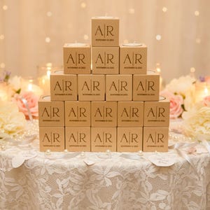 May include: A pyramid of personalized candle boxes with the initials "AR" and the date "November 16, 2023" printed on them. The boxes are arranged on a lace-covered table, surrounded by floral arrangements of pink and white roses, and pearls.