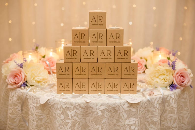 May include: A pyramid of personalized candle boxes with the initials "AR" and the date "November 16, 2023" printed on them. The boxes are arranged on a lace-covered table, surrounded by floral arrangements of pink and white roses, and pearls.