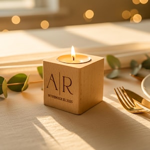 May include: A lit candle in a wooden block with the initials "AR" and the date "November 10, 2025" engraved on the front. The candle sits on a table with gold silverware, a white plate, and greenery.