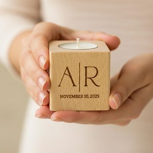 May include: A square wooden candle holder with a lit white candle. The holder is engraved with the initials "AR" and the date "NOVEMBER 10, 2025". The candle holder is held in two hands.