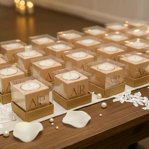 May include: A collection of wooden candle holders with clear plastic tops, each containing a white tea light. The wooden blocks are engraved with initials and sit on gold-colored bases. The arrangement is on a wooden table with decorative elements.