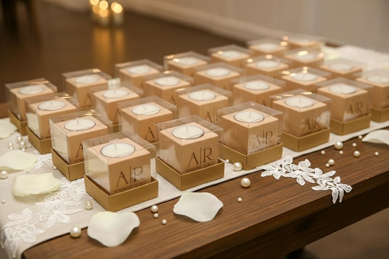 May include: A collection of wooden candle holders with clear plastic tops, each containing a white tea light. The wooden blocks are engraved with initials and sit on gold-colored bases. The arrangement is on a wooden table with decorative elements.