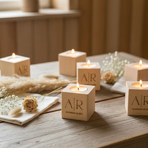 May include: Wooden cube candle holders with lit candles, engraved with initials and a date. The holders are arranged on a wooden table with dried flowers and a white napkin, creating a warm, rustic ambiance.