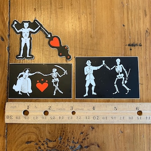 May include: Four black and white pirate flag stickers with a red heart design. The stickers feature a skeleton holding a sword, a skeleton holding a cup, a skeleton dancing with a woman, and a skeleton holding a sword and aiming it at a heart.