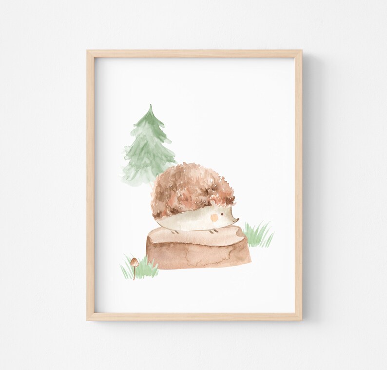 May include: Watercolor illustration of a hedgehog sitting on a tree stump, with a green tree and grass in the background. The artwork is framed in a light wood frame. The hedgehog is brown and white, and the overall theme is nature.