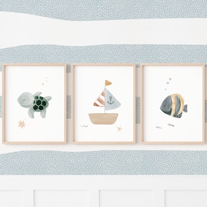 Op de afbeelding: Drie ingelijste aquarelprints met een nautisch thema. De prints tonen een schildpad, een zeilboot en een vis. Het kunstwerk is in zachte kleuren, met een licht houten frame, en is geplaatst tegen een blauw-wit gestreepte achtergrond.