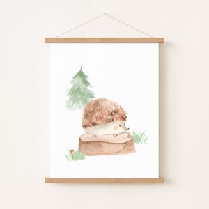 May include: Watercolor print of a hedgehog sitting on a log, with a green tree and grass in the background. The hedgehog has brown and beige fur. The artwork is displayed on a white background and framed with a wooden hanger.