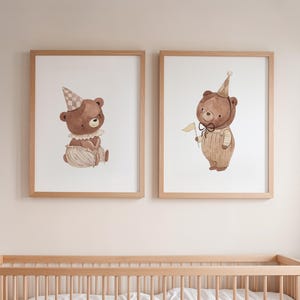 Könnte beinhalten: Zwei gerahmte Aquarelldrucke mit braunen Teddybären mit Partyhüten. Ein Bär sitzt und trägt einen Rüschenkragen und eine Hose. Der andere Bär steht und hält eine kleine Fahne und trägt eine Latzhose. Die Rahmen sind aus hellem Holz.