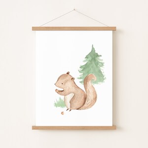 May include: Watercolor print of a brown squirrel holding a nut, with a green pine tree in the background. The artwork is displayed on a white background and framed with a wooden magnetic poster hanger. The style is whimsical and suitable for a nursery.