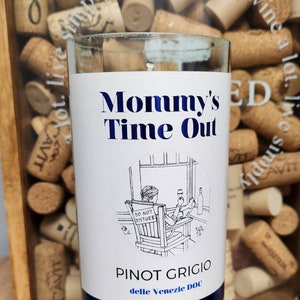 May include: A white candle in a clear glass bottle with a blue label that says "Mommy's Time Out" and "Pinot Grigio delle Venezie DOC". The label also features a cartoon illustration of a person sitting in a chair with a "Do Not Disturb" sign.