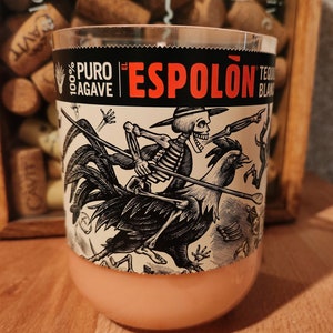 May include: A clear glass with a peach-colored base and a black and white label. The label features the text "100% PURO AGAVE" and "EL ESPOLÓN TEQUILA BLANCO". A skeleton figure rides a rooster in the illustration.