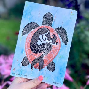 May include: A notebook with a mermaid illustration inside a turtle shell design. The mermaid has long flowing hair and a fish tail. The turtle shell is orange, and the background is blue with watercolor effects. The words "companion ship" and "sophie y" are written on the notebook.