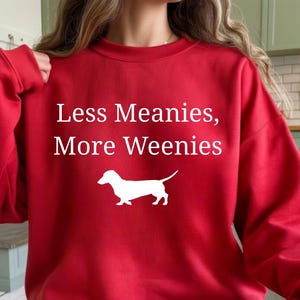 May include: A red sweatshirt with the text "Less Meanies, More Weenies" in white. A white silhouette of a dachshund dog is below the text. The sweatshirt is a casual style.