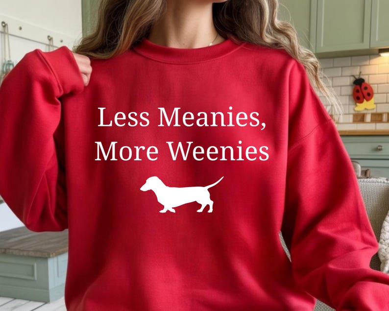 May include: A red sweatshirt with the text "Less Meanies, More Weenies" in white. A white silhouette of a dachshund dog is below the text. The sweatshirt is a casual style.