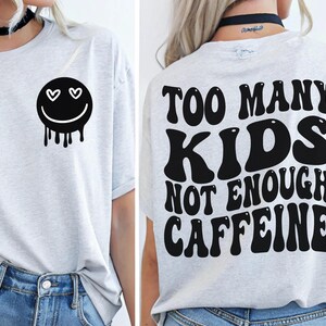 Könnte beinhalten: Hellgraues T-Shirt mit einem schwarzen Smiley-Gesicht-Motiv auf der Vorderseite, mit Herzaugen und tropfenden Details. Auf der Rückseite des Shirts steht der Text "TOO MANY KIDS NOT ENOUGH CAFFEINE" in einer kräftigen Retro-Schrift. Ein lässiges T-Shirt für den Alltag.