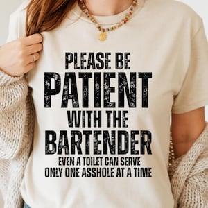Op de afbeelding: Een beige t-shirt met de tekst "PLEASE BE PATIENT WITH THE BARTENDER" in grote, zwarte letters. Daaronder staat in kleinere letters: "EVEN A TOILET CAN SERVE ONLY ONE ASSHOLE AT A TIME."