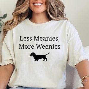 May include: Cream-colored t-shirt with the text "Less Meanies, More Weenies" and a black silhouette of a dachshund. The shirt is a casual style.