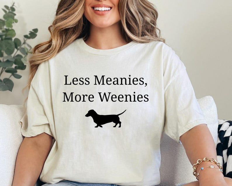 May include: Cream-colored t-shirt with the text "Less Meanies, More Weenies" and a black silhouette of a dachshund. The shirt is a casual style.