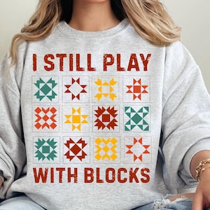 May include: A grey sweatshirt with the text "I Still Play With Blocks" in red and black. The text is surrounded by nine colourful quilt blocks.