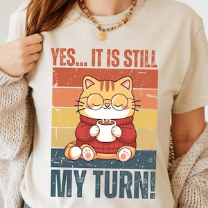 Puede incluir: Camiseta beige con un gato de dibujos animados con gafas y un suéter rojo, sosteniendo una taza de café. El texto "YES... IT IS STILL MY TURN!" está impreso encima y debajo de la ilustración del gato, sobre un fondo retro.