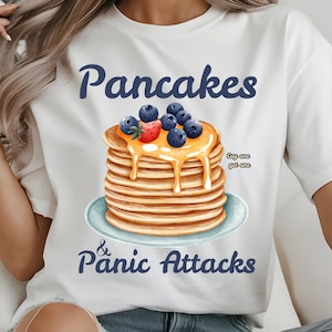 Könnte beinhalten: Weißes T-Shirt mit einem Grafikdruck von einem Stapel Pfannkuchen, der mit Blaubeeren und Erdbeeren belegt und mit Honig beträufelt ist. Der Text "Pancakes & Panic Attacks" ist unter dem Bild gedruckt. Der Text "Buy one, get one" ist in kleinerer Schrift über den Pfannkuchen gedruckt.