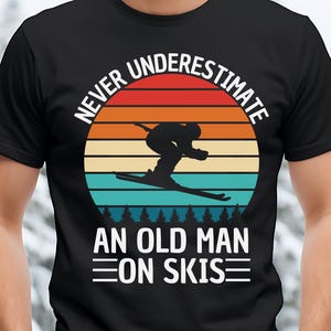 Op de afbeelding: Zwart t-shirt met een retro design met een silhouet van een skiër tegen een zonsondergang. De tekst luidt "NEVER UNDERESTIMATE AN OLD MAN ON SKIS" in het wit.