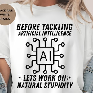 Op de afbeelding: Wit T-shirt met een zwart grafisch ontwerp. Het ontwerp bevat de tekst "BEFORE TACKLING ARTIFICIAL INTELLIGENCE" boven een printplaat met "AI" in het midden en "LETS WORK ON NATURAL STUPIDITY" eronder.