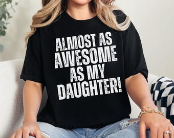 May include: Black t-shirt with white text that reads "Almost as awesome as my daughter!"