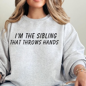 Op de afbeelding: Een grijs sweatshirt met de tekst "I'm the sibling that throws hands" gedrukt op de voorkant.
