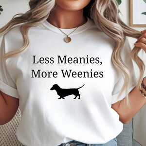 May include: White t-shirt with the text "Less Meanies, More Weenies" in black, along with a black silhouette of a dachshund. The shirt is a casual style.