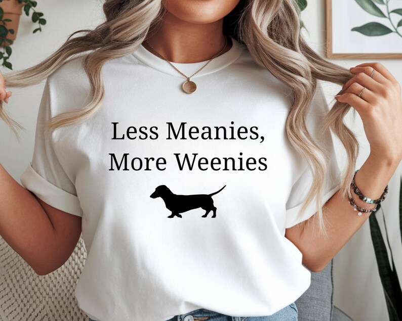 May include: White t-shirt with the text "Less Meanies, More Weenies" in black, along with a black silhouette of a dachshund. The shirt is a casual style.