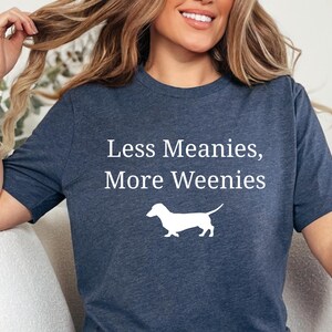 May include: A navy blue t-shirt with the text "Less Meanies, More Weenies" and a white silhouette of a dachshund. The shirt is made of a soft material.
