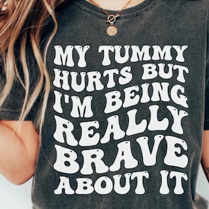 May include: A dark grey t-shirt with white text that reads "My tummy hurts but I'm being really brave about it". The text is in a retro, bubble-like font.