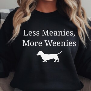 May include: Black sweatshirt with the text "Less Meanies, More Weenies" in white, along with a white silhouette of a dachshund dog. The sweatshirt is a casual style.