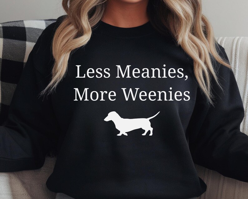 May include: Black sweatshirt with the text "Less Meanies, More Weenies" in white, along with a white silhouette of a dachshund dog. The sweatshirt is a casual style.